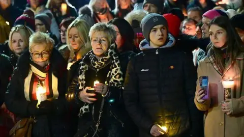 Pacemaker Crowds of people at a vigil in Banbridge. Some at the front are holding candles. At the front are three women and a young man. Two of the women have blonde hair and are wearing colourful scarves. The young man is wearing a dark jacket and a dark hat. The other woman has dark hair and a light coat.