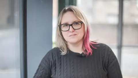 Image of Dr Lynsay Shepherd. It's a head and shoulders shot and she is staring straight at the camera. She has shoulder length blonde hair with pink highlights. She's wearing black-rimmed glasses and black knitted jumper.