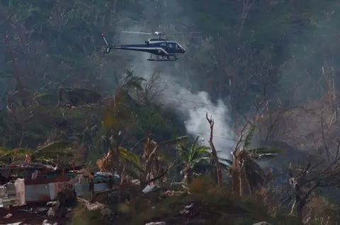 Gonzalo Fuentes/Reuters A helicopter from the French Gendarmerie flies over the Longoni village in the aftermath of Cyclone Chido in Mayotte. Trees and buildings are flattened or badly damaged and smoke rises from the ground, from where two people inspect the damage.
