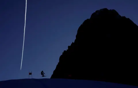 Leonhard Foeger/Reuters Skier Kyle Negomir is silhouetted against the landscape and blue sky as he practises a ski run at Val Gardena, in Italy. He appears tiny beside a giant rock, which is also in silhouette, while a white vapour trail stands in the sky behind them both. 