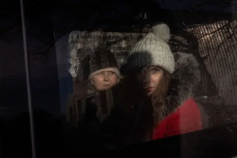 Roman Pilipey/AFP Two women, both wearing hats and warm coats, look out from the window of a car