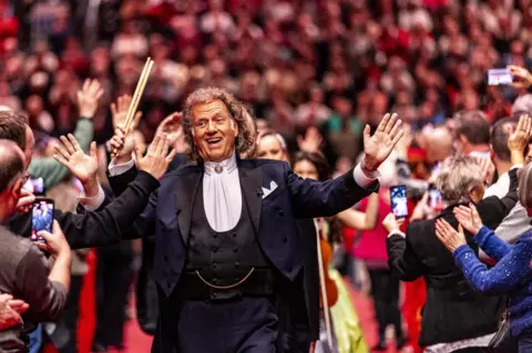 Marcel van Hoorn/EPA-EFE Violinist Andre Rieu, dressed in a black suit with white shirt, walks, smiling, arms wide apart, through a crowd of people, some taking photographs with mobile phones, in Maastricht, in the Netherlands.
