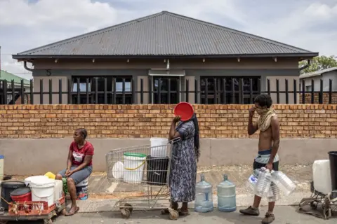 Roberta Ciuccio/AFP People queue for water in Johannesburg. One person is sitting at the roadside with a number of containers, another stands, holding on to a shopping trolley containing vessels and also covering her face; a third stands with a number of large containers in his hand.