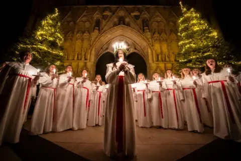 Danny Lawson/PA Media A young woman wearing a crown of lighted candles and holding another candle with both hands, sings surrounded by other members of the female choir, who are also holding candles. Behind them at York Minster are two Christmas trees, decorated with lights.