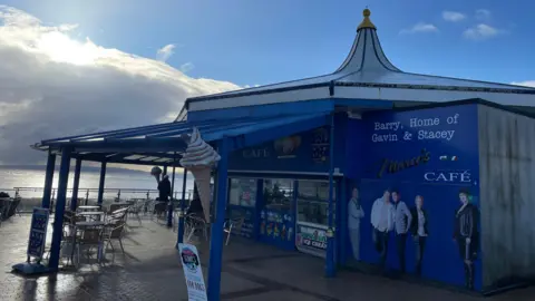 Marco's Cafe on Barry Island. The cafe is a bold blue with outdoor seating overlooking the sea. On the wall there are life-sized images on Nessa, Uncle Bryn, Smithy, Gavin and Stacey.