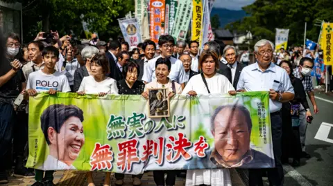 Getty Images A crowd of people walks down the street holding a banner showing the faces of Iwao Hakamata and Hideko Hakamata 