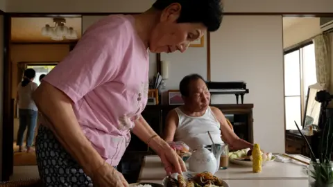 A woman in a pink shirt puts meals down on a kitchen table while a man in a white singlet sits at the head of the table