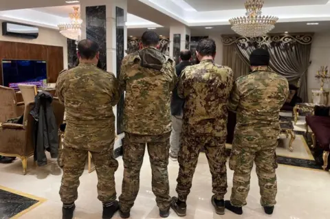 four men in camo gear stand facing away from the camera