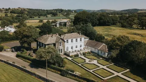 Christie & Co An aerial shot of the old grammar school building in Cartmel. The large white building is L-shaped and sits next to a stone-clad one. There is a large manicured garden at the front.