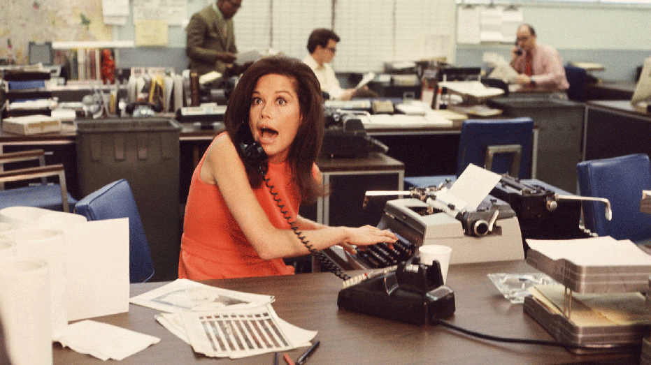 American actress Mary Tyler Moore mouths surprise on the telephone while simultaneously typing as others work in the background in a scene from 'The Mary Tyler Moore Show' (also known as 'Mary Tyler Moore'), Los Angeles, California, early 1970s. Moore wears a sleevless orange dress as she sits behind her desk. (Photo by CBS Photo Archive/Getty Images)