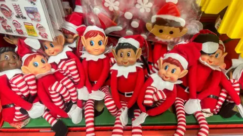 Getty Images A shelf full of Elf on the Shelf toys. The elves are dressed in a red long sleeved top with red and white striped bottoms. 