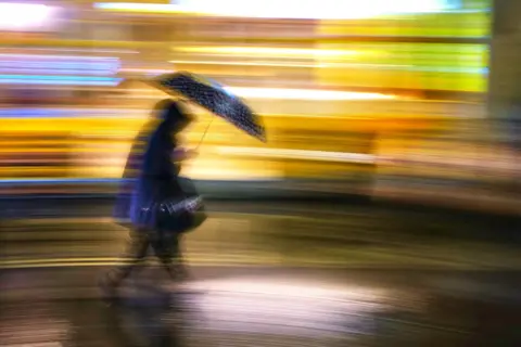 John Barnes A person with an umbrella passes bright lights. The person has their head down and is walking against the rain.