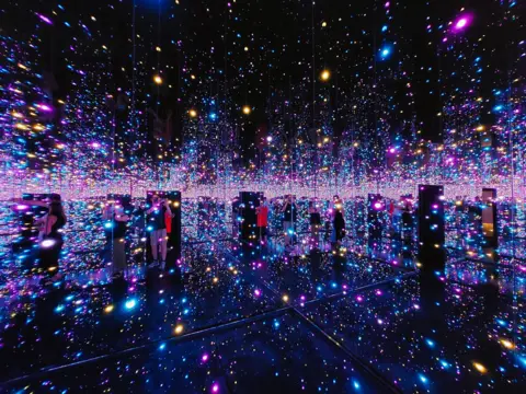 Pokman Cheung Bright lights in an infinity room. The room is dark and the speckled lights are shades of purple and blue.