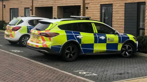 Alex Stevanovic/BBC Two police cars are parked outside a modern set of flats, which have cream coloured bricks and windows with black rims.