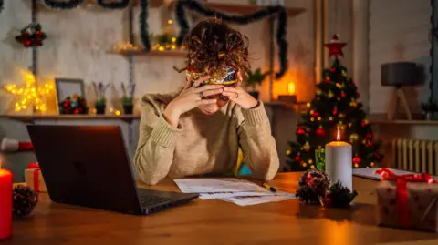 Getty Images A woman has her hands resting on her forehead as she looks at bills on an oak table and a black laptop next to her. She has curly brown hair tied up on top of her head and is wearing a beige jumper. In the background, there is a Christmas tree and tinsel adorning shelves.