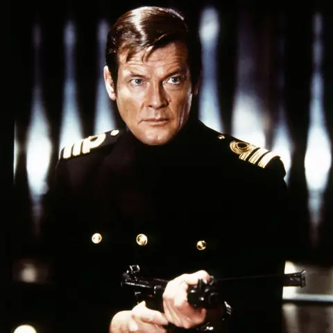 Getty Images Sir Roger Moore is seen on the set of the James Bond film, The Spy Who Loved Me. He is wearing a double-breasted naval officer's uniform and is pointing a gun at someone off camera. He has a serious and purposeful expression on his face. 