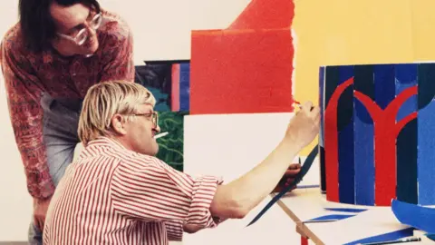 © Sabina Sarnitz. Courtesy Luna Luna, LLC A young David Hockney smokes a cigarette as he paints a blue and red structure with someone peering over his shoulder. 