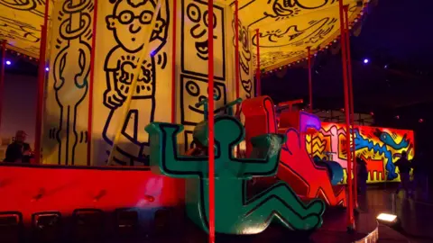 BBC/Ottilie Mitchell Keith Haring's carousel with bold cartoon figures is in close up.