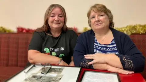 Two middle-aged women sit side by side on a club sofa with framed photographs and documents on a table in front of them. Julie, to the left, has long brown hair and wears a green Christmas T-shirt. Janet, to the right, has short, light-brown hair and wears a blue and white patterned top and a dark blue cardigan. 