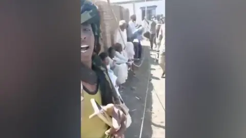 Social media Survivors are seen sitting against a wall while a guard films them. 