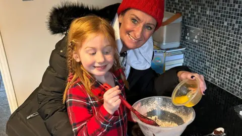 Susannah Morgan Susannah is wearing a red wooly hat and fluffy hooded coat and is smiling at the camera. She is baking a cake with her daughter who is stirring a red spoon in a bowl. Her daughter has long ginger hair and is wearing a red and black checked shirt.