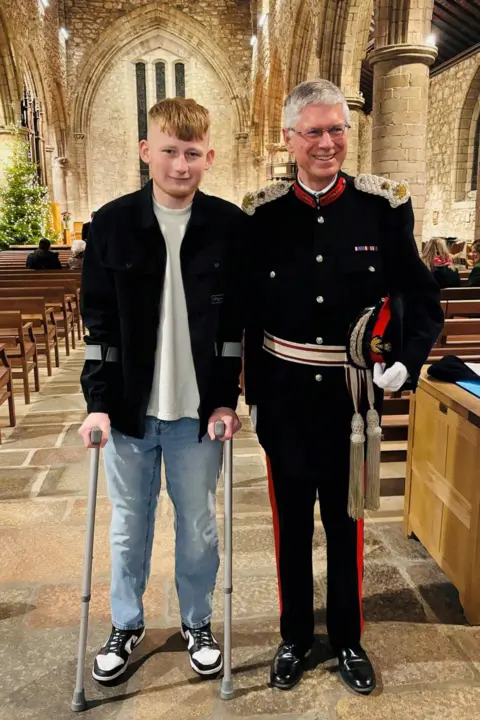 Golebiewski family Teenage boy with ginger hair wearing light blue jeans, a light T-shirt and black casual jacket and trainers, standing on crutches, next to an older man with grey hair, smiling, wearing a dark Lord Lieutenant uniform, both standing in the aisle of a cathedral.