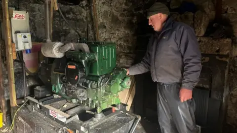 The image shows a man looking at a diesel generator. He is an older man wearing waterproofs with a blue jacket  