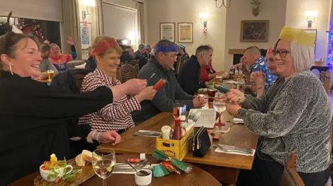 Shona Anderson and Sam Wood are in the foreground of the picture, wearing party hats in a room full of lots of other people having a Christmas party  