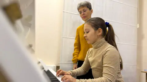 BBC/Kostas Kallergis Anna playing the piano, watched by Viktoria