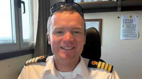 BAS Capt Will Whatley is smiling at the camera. He has fair hair and sunglasses balanced on his head. He is wearing a white shirt with epaulettes. He is sitting in a chair on board a ship