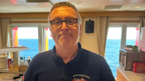 BAS Nick Greenwood is looking at the camera and has short fair hair and is wearing spectacles. He has a black polo-shirt on with the insignia of the British Antarctic Survey ship on the chest. He is standing in a canteen area on a boat. Food can be seen to one side and the blue sea is in the background