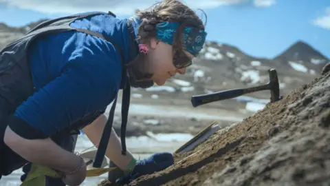 Sam Hunt Rowan  Whittle is seen in a side view, wearing a blue bandana around her head. She has dark hair, worn in plaits and is wearing dark sunglasses and a blue top. She is looking closely at some rocks and have various tools with her
