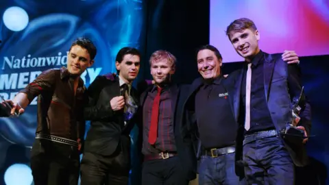 PA Media Host Jools Holland congratulates Franz Ferdinand on winning the Mercury Music Prize in 2004. The band and Holland have their arms wrapped around each other while singer Alex Kapranos, on the far right, holds the award in his hand. The band are mostly dressed suits with ties, except for guitarist Nick McCarthy who is dressed casually.