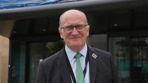 Nikki Fox/BBC Nick Hulme smiles at the camera standing outside a hospital building. He is largely bald and is wearing glasses. He wears a black suit with a light blue shirt underneath and green tie. He wears a Pride flag pin badge on his chest.