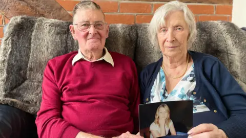 Bryson and Margaret Garlick sitting together on a sofa at their home holding a photo of their daughter