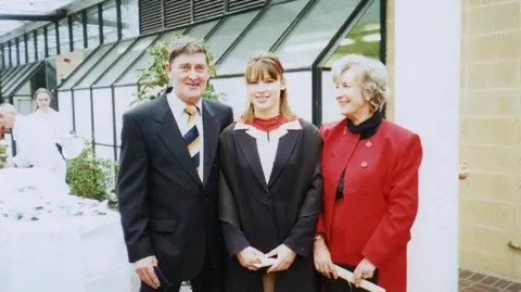 Family Bryson and Margaret Garlick pictured with their daughter Deborah on her graduation day in 1996. Deborah is in a gown, her mother is looking at her smiling. Her father is smiling into the camera.