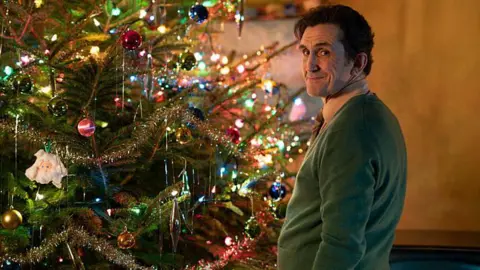 BBC/Neal Street Productions Stephen McGann as Dr Turner, standing by a brightly decorated Christmas tree in a green jumper