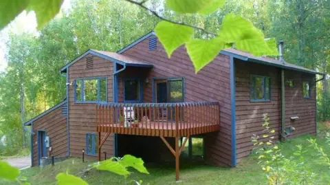 Liz Barlow A wooden house with a deck on stilts. It is surrounded by hedges and trees. A small path leads away from the house.