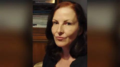 Debbie Foster A woman with dark red hair, wearing a black cardigan over a read and black striped top looks into camera as she sits in a dimly lit room.