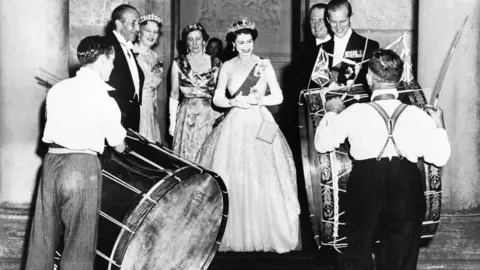 Getty Images A black and white archive photo showing Queen Elizabeth II in a formal ballgown standing in the centre of the photo. In front of her are two men in white shirts playing Lambeg drums. To her left is her husband Prince Philip who is wearing white tie. 