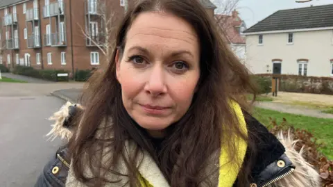 Sam Read/BBC Sophie Pritchard stands on the estate. She is looking at the camera with a solemn expression. She is wearing a padded blue coat, bright green, beige and brown scarf, and has long brown hair and brown eyes. 