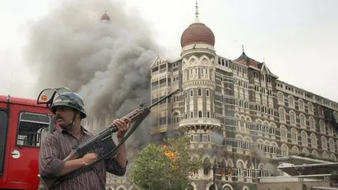 Getty Images An Indian security official wearing a red striped shirt and a helmet is holding a gun as he stands alert as smoke and flames billow out from a section of The Taj Mahal hotel in Mumbai on November 29, 2008