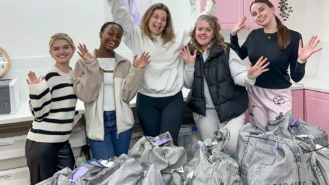 Isobel Perl Isobel and four other young women with their hands in the air, behind lots of parcels