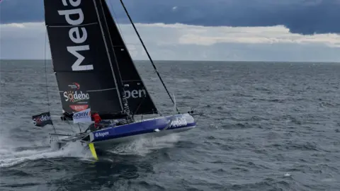 James Tomlinson A blue sailing boat with a large black sail is travelling through water at speed. The sky is grey.