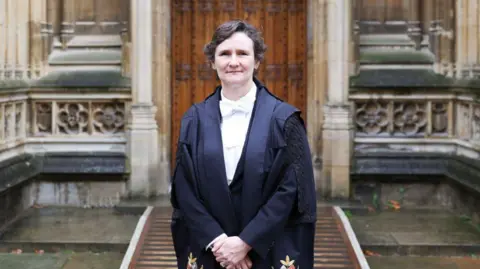 University of Oxford Prof Tracey in black gowns and a white shirt. She is stood with her arms down in front and has short brown hari. Behind her is a rather old looking stone doorway.