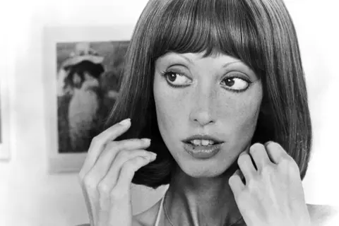 Getty Images Black and white head shot of Shelley Duvall, taken from the 1977 film Three Women. She is looking away from the camera, wearing make-up and touching her hair, which is shoulder-length.