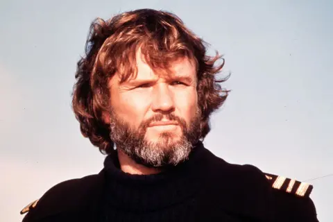 Getty Images Film still of Kris Kristofferson, taken in the 1970s against a background of sky - he has reddish hair, a greying beard and is wearing a black turtle-neck sweater with epaulettes