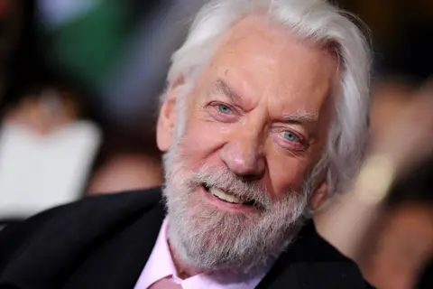 Getty Images Close-up photo of Donald Sutherland pictured at the premiere of The Hunger Games: Catching Fire in 2013, when he was 78. He is grey-haired and bearded and is smiling. He wears a black jacket and a pink shirt and tie.