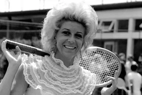 Getty Images A black and white photo of Lea Pericoli holding a tennis racket behind her shoulders. She is wearing bold earrings and a very frilly tennis top.