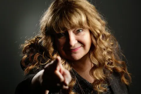 Alamy Publicity shot of Janey Godley pointing straight at the camera and smiling. She has long auburn hair, slightly curly and is wearing a black top with silver flecks